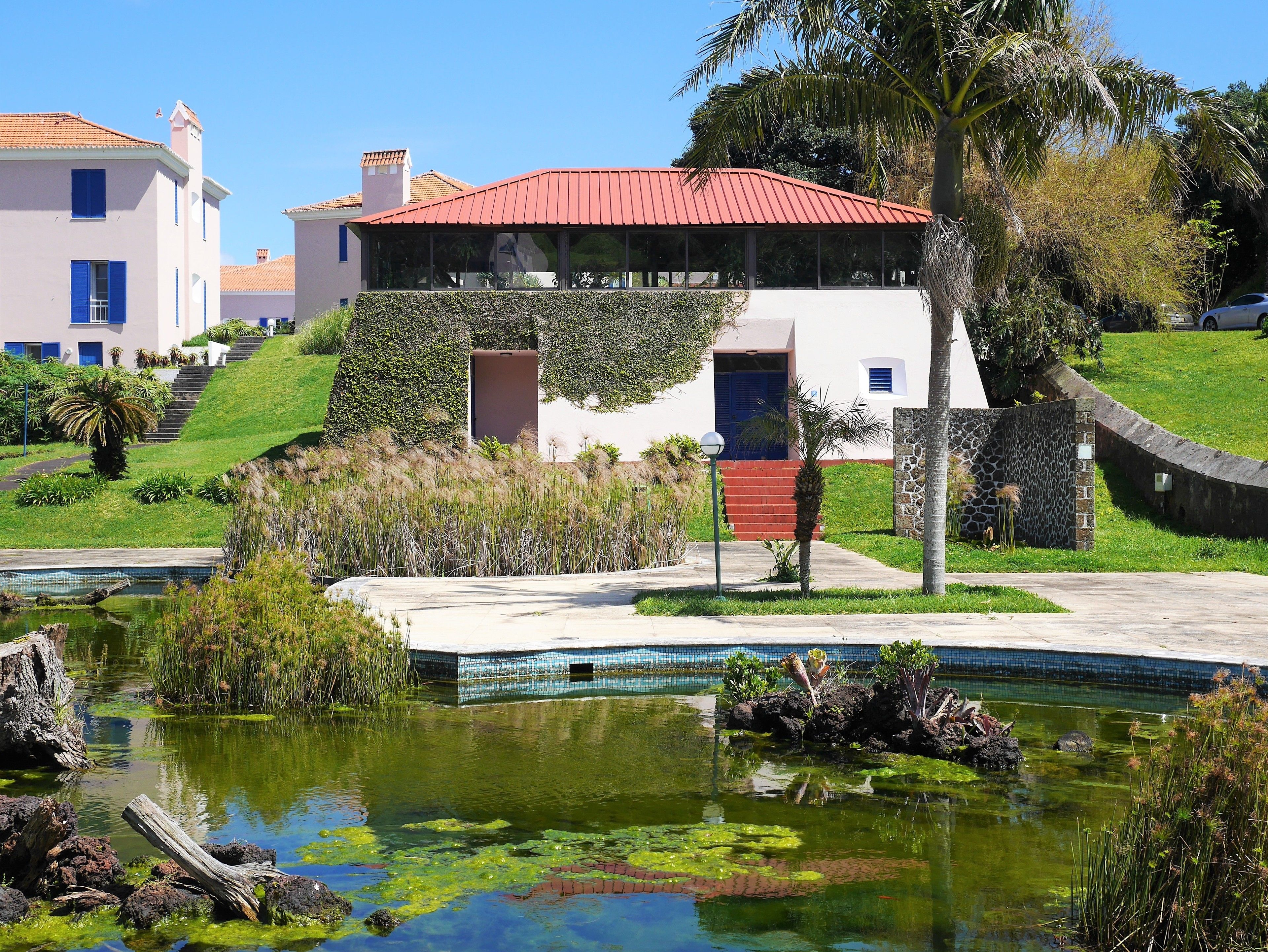 Un edificio moderno con techo rojo, piscina con lirios acuáticos y un jardín bien cuidado.