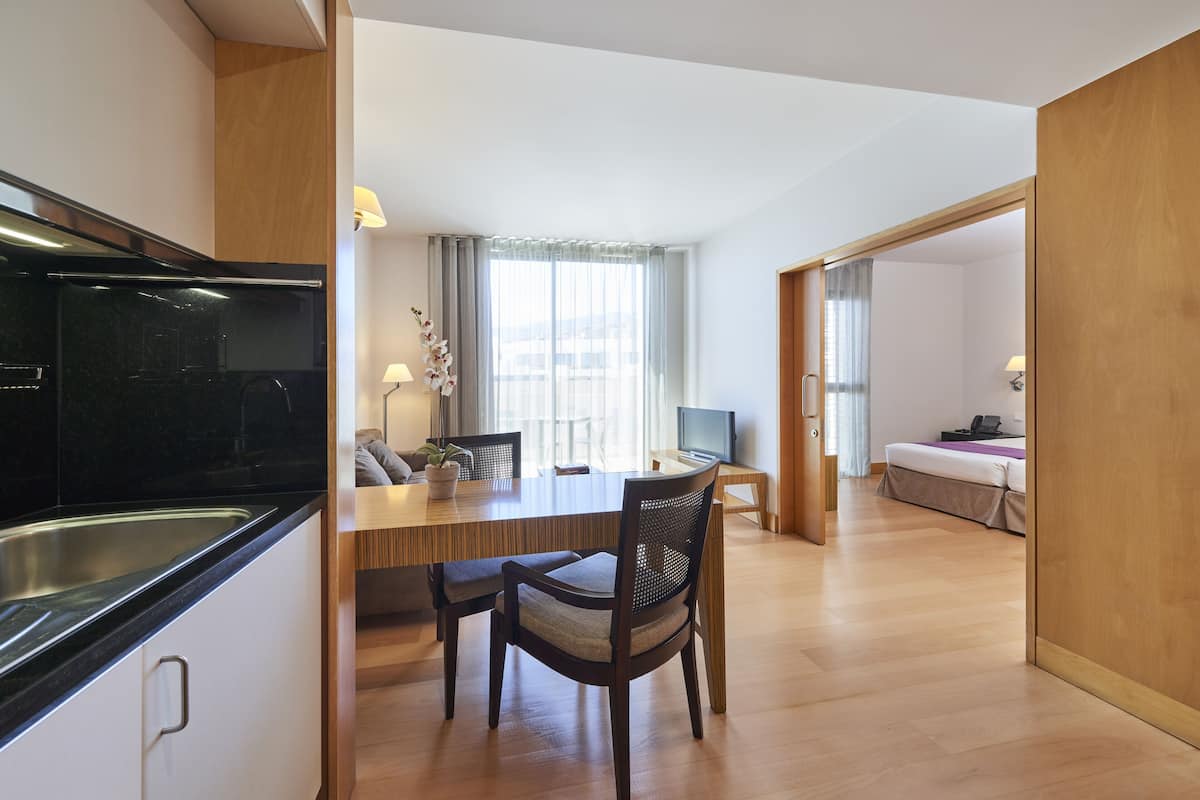 A modern kitchen with a black countertop and stainless steel sink, a dining area with a wooden table and chairs, and a bedroom with a bed and a TV.