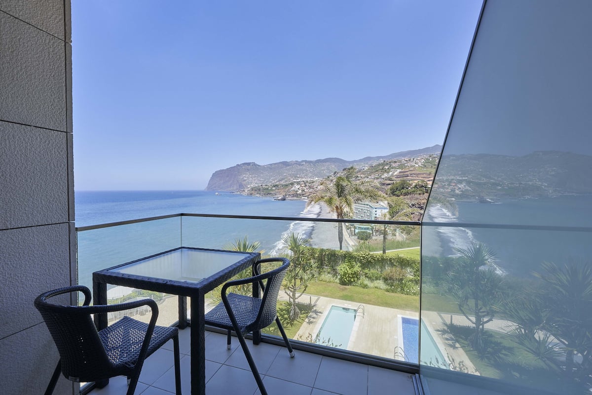 A balcony with a glass table and chairs overlooking a coastal view with a swimming pool.
