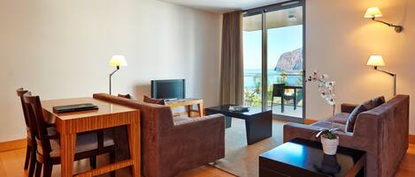 Une chambre d’hôtel moderne avec une table à manger en bois, deux canapés, une petite télévision et un balcon offrant une vue sur la mer.