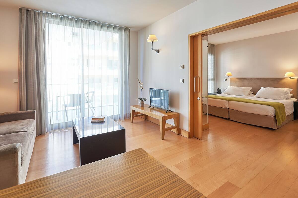 A hotel room with a bed, a desk with a TV, a sofa, and a dining table.
