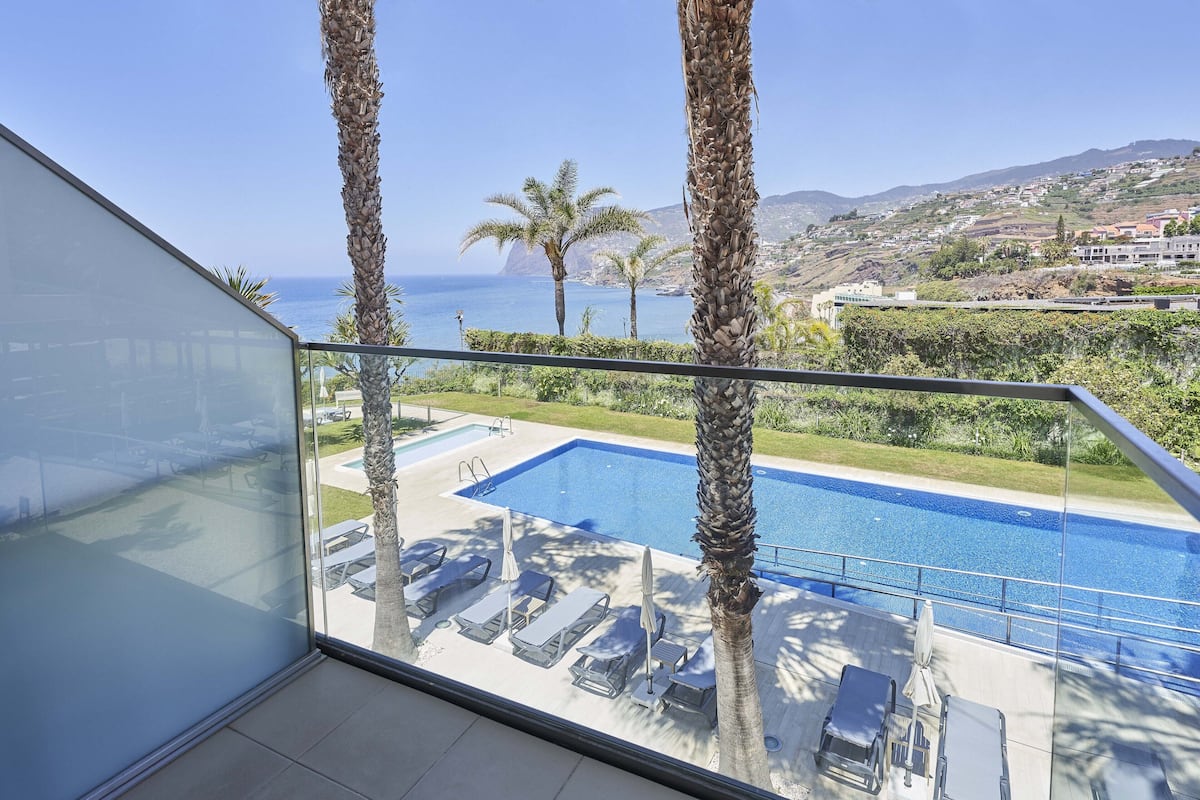 A balcony with a view of a swimming pool, palm trees, and a distant coastline.