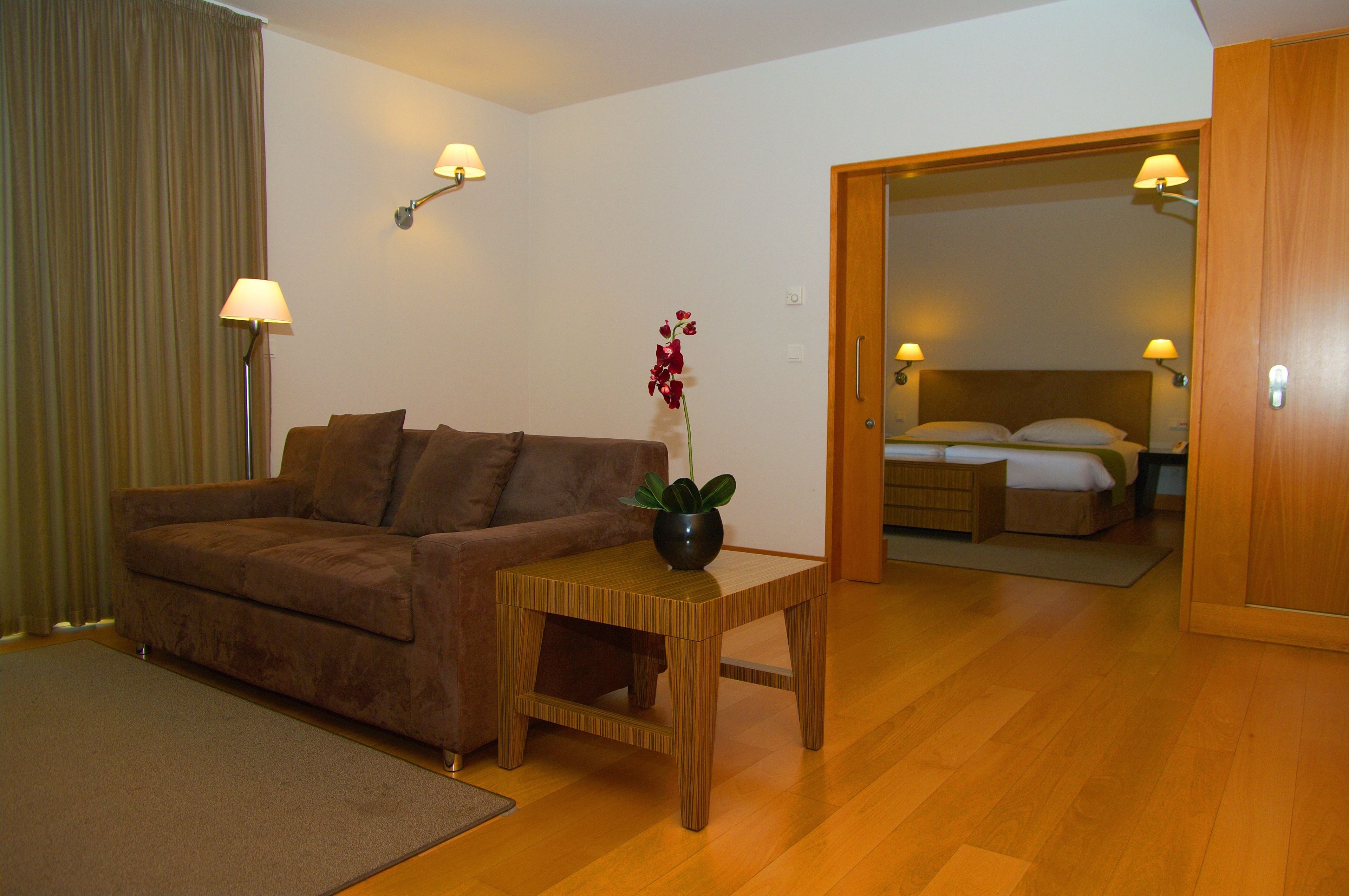 Quarto de hotel com sofá marrom, uma mesa lateral de madeira com planta em vaso, e uma cama com cabeceira de madeira.