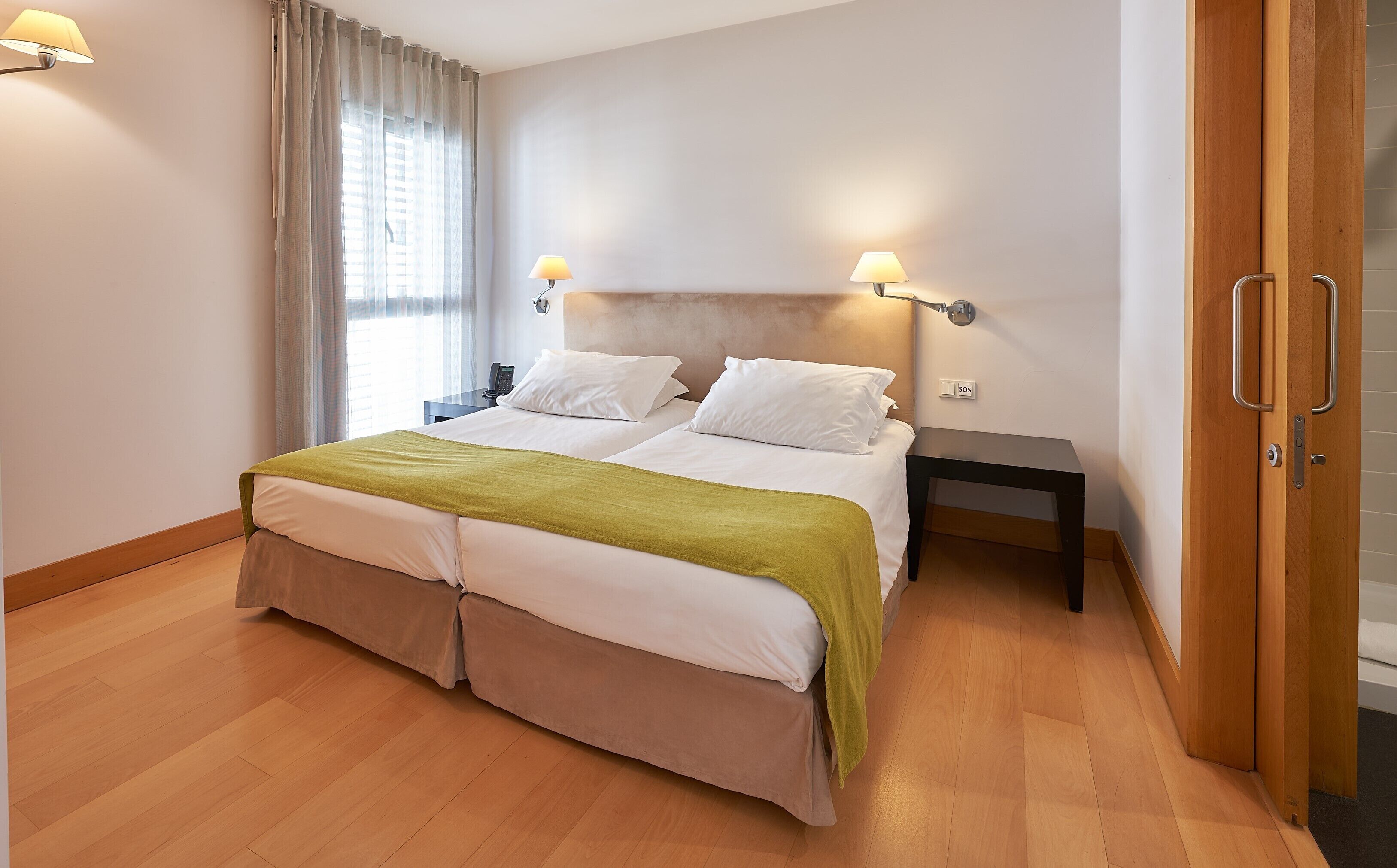 Een tweepersoonsbed met een groene plaid, nachtkastjes met lampen, een houten deur en een raam met gordijnen.