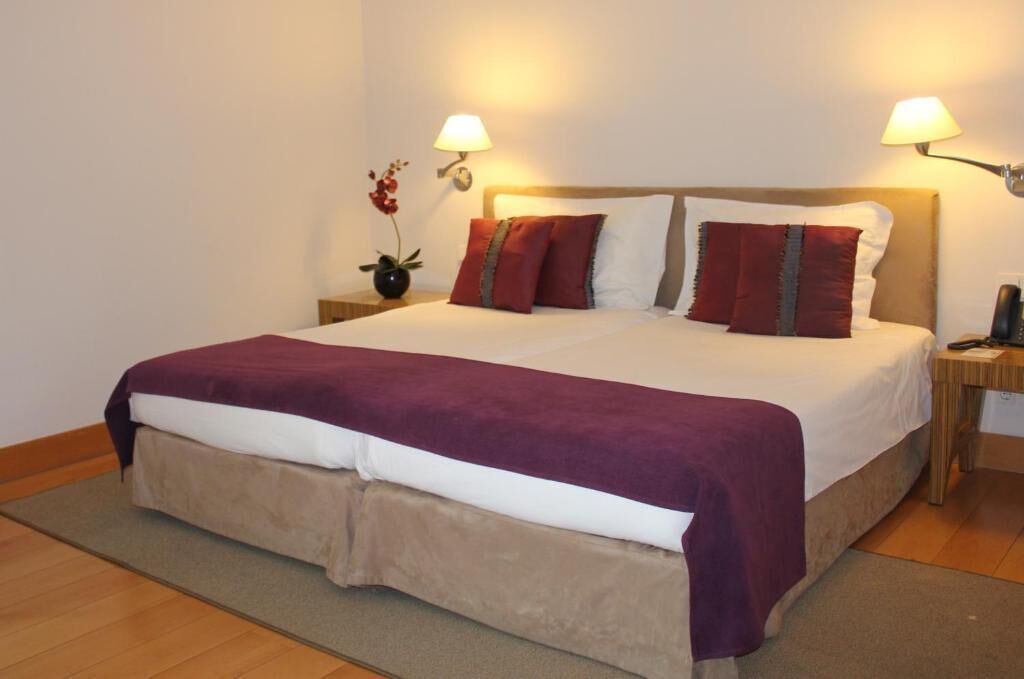 Una cama bien hecha con una colcha blanca y morada, dos almohadas rojas y una base de cama color beige con falda. Hay dos mesitas de noche con lámparas y un jarrón pequeño con flores.