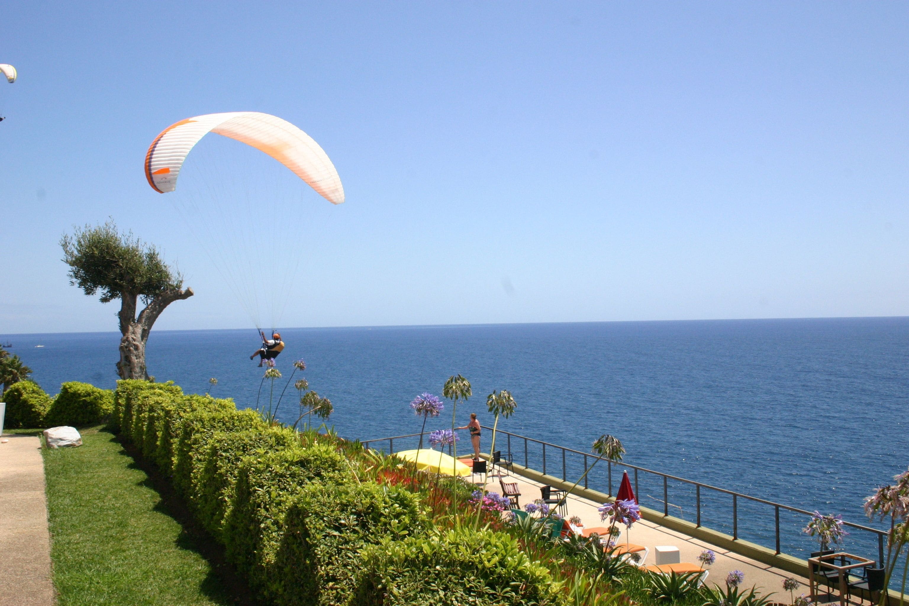 Eine Küsten-Terrasse mit Blick auf das Meer, ein Parasegel in der Ferne und eine Person am Geländer.