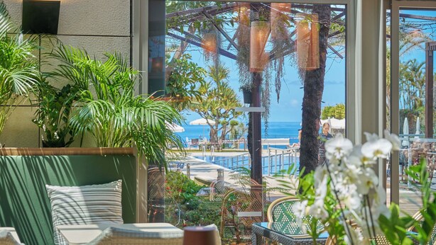 A sunlit indoor seating area with a view of the ocean, featuring a sofa, chairs, and a table. - Golden Residence Hotel (Funchal)