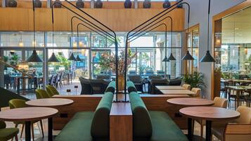 Een modern café met ronde tafels, zitplaatsen met groene kussens en grote ramen met uitzicht op het terras.