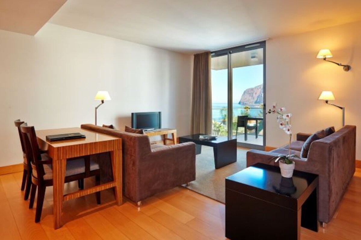 Une chambre d’hôtel moderne avec une table à manger en bois, deux canapés, un téléviseur et un balcon avec vue.