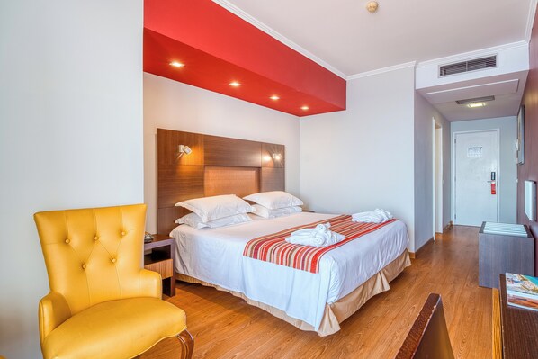 Une chambre d’hôtel avec un lit, un fauteuil jaune, une tête de lit en bois et un mur d’accent rouge.