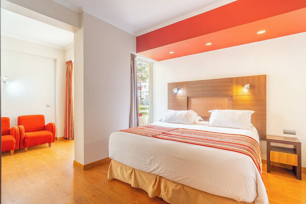 A modern hotel room with a large bed, a wooden headboard, a small bedside table, and a bright red accent wall.