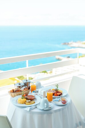 Une table de petit-déjeuner avec vue sur l’océan, une nappe blanche et divers aliments pour le petit-déjeuner, notamment des œufs, des toasts et du jus d’orange.
