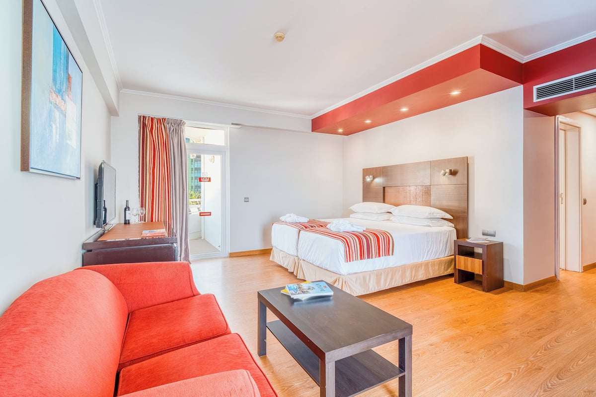 A modern hotel room with a large bed, a red sofa, a small table, and a TV on a stand.