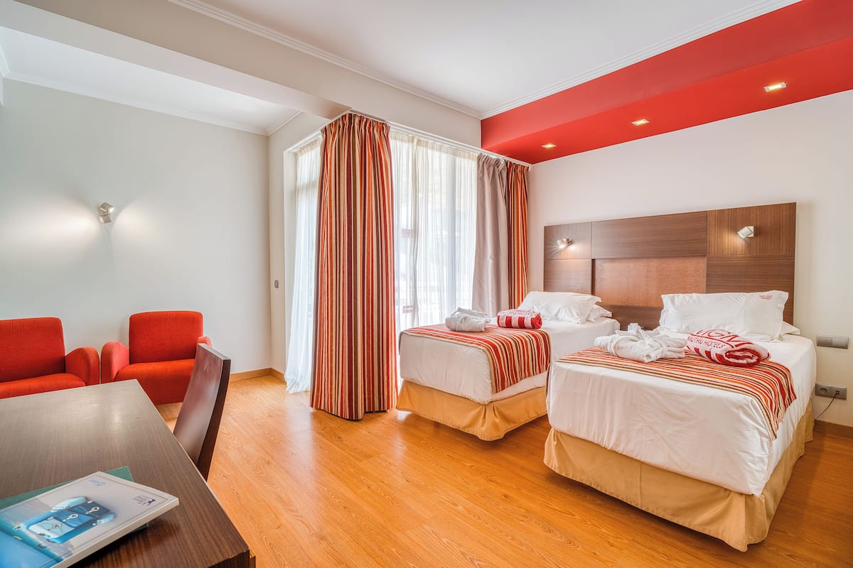 A hotel room with two beds, a red sofa, a wooden table, and a window with curtains.