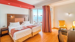 A hotel room with two beds, a wooden floor, a red accent wall, a large window with curtains, and a view of the sea. - Muthu Raga Madeira Hotel (Funchal)