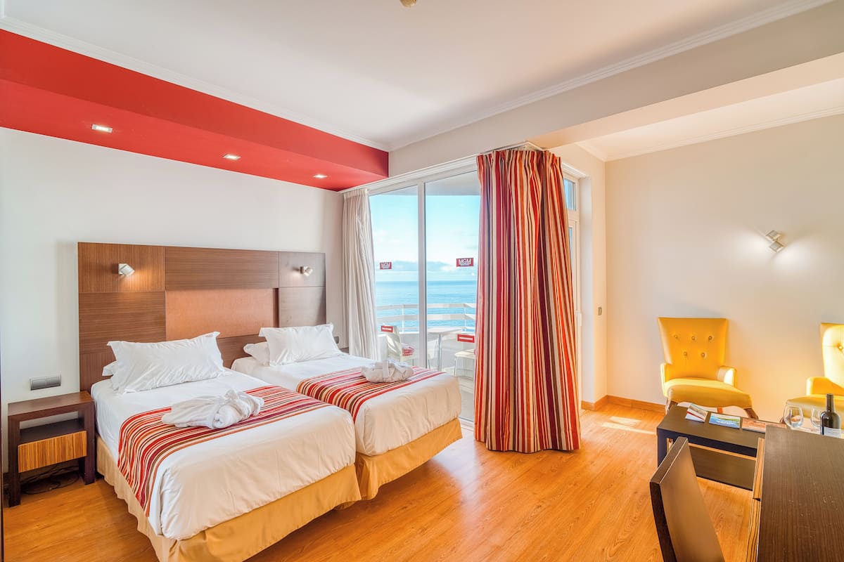 A hotel room with two beds, a wooden floor, a red accent wall, a large window with curtains, and a view of the sea.