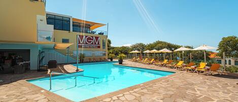 A swimming pool area with lounge chairs and umbrellas, a building with a 'MGM' sign, and a stone-tiled deck.