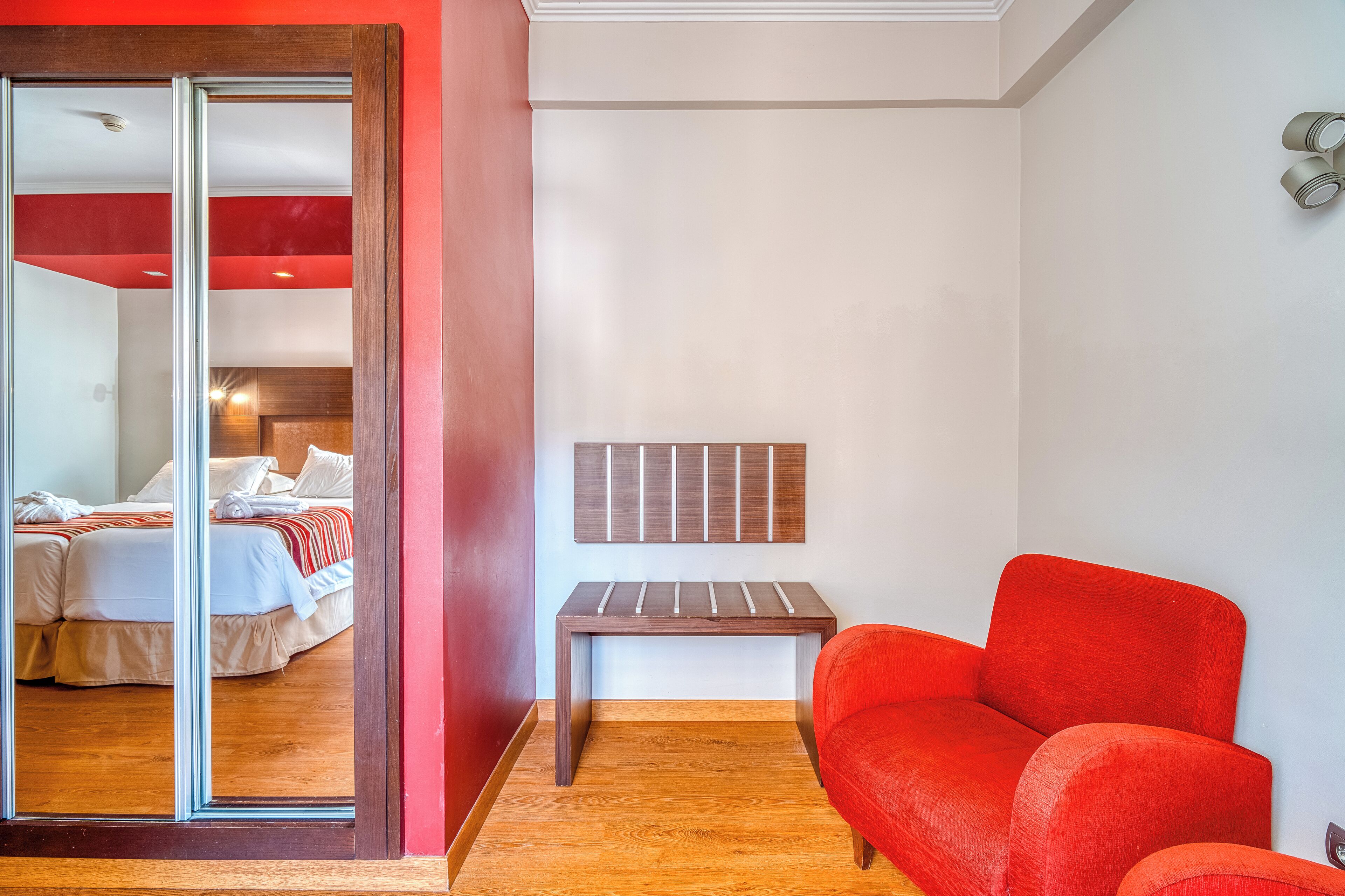 A hotel room with a sliding glass door leading to a bedroom, a red sofa, a small wooden table, and a wall-mounted light fixture.
