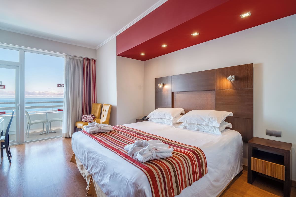 A hotel room with a large bed, a wooden nightstand, a chair, and a view of the sea through a sliding glass door.