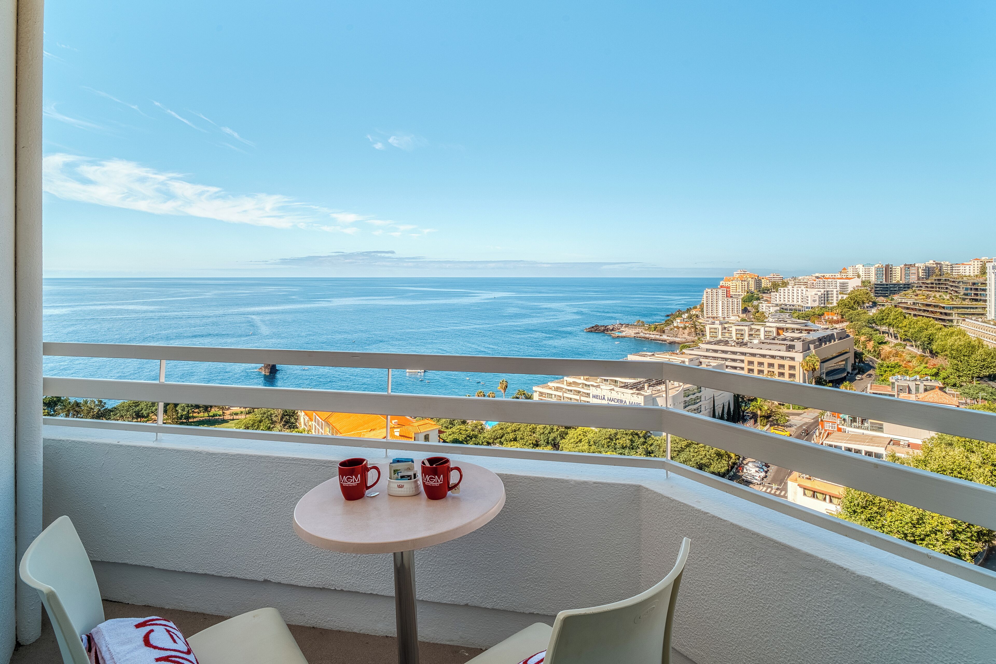 A balcony with a table and chairs, overlooking a coastal cityscape with buildings and the sea.