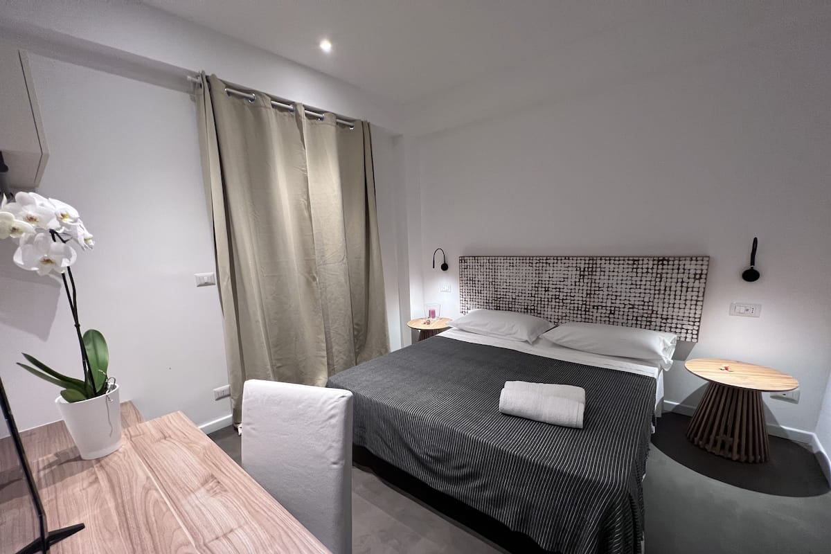 Moderni hotellihuone, jossa on sänky, yöpöydät, työpöytä ja ruukussa oleva kasvi.