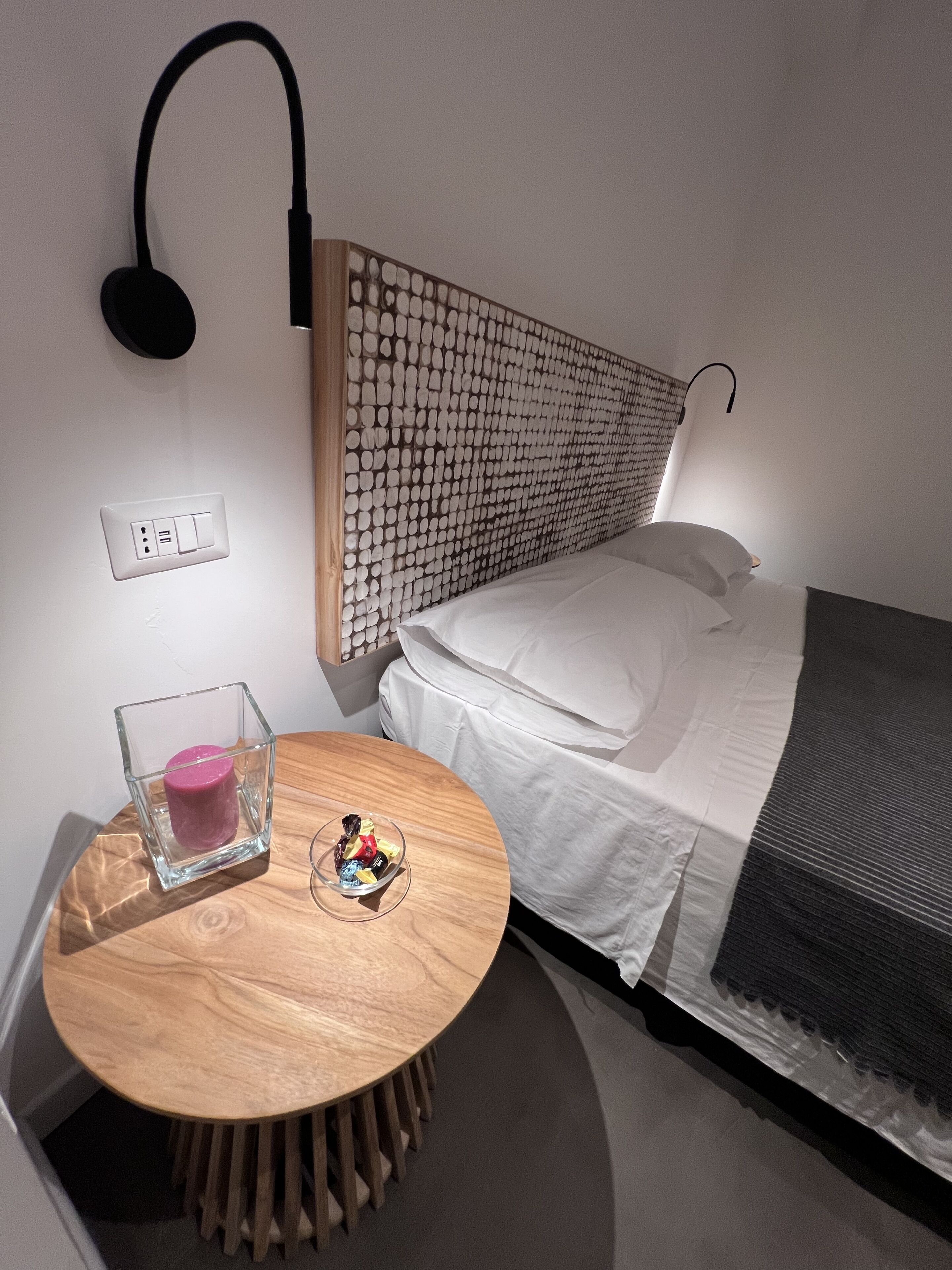 A modern bedroom with a wooden headboard, a round wooden table with a glass candle holder, a small bowl with items, and a wall-mounted lamp.