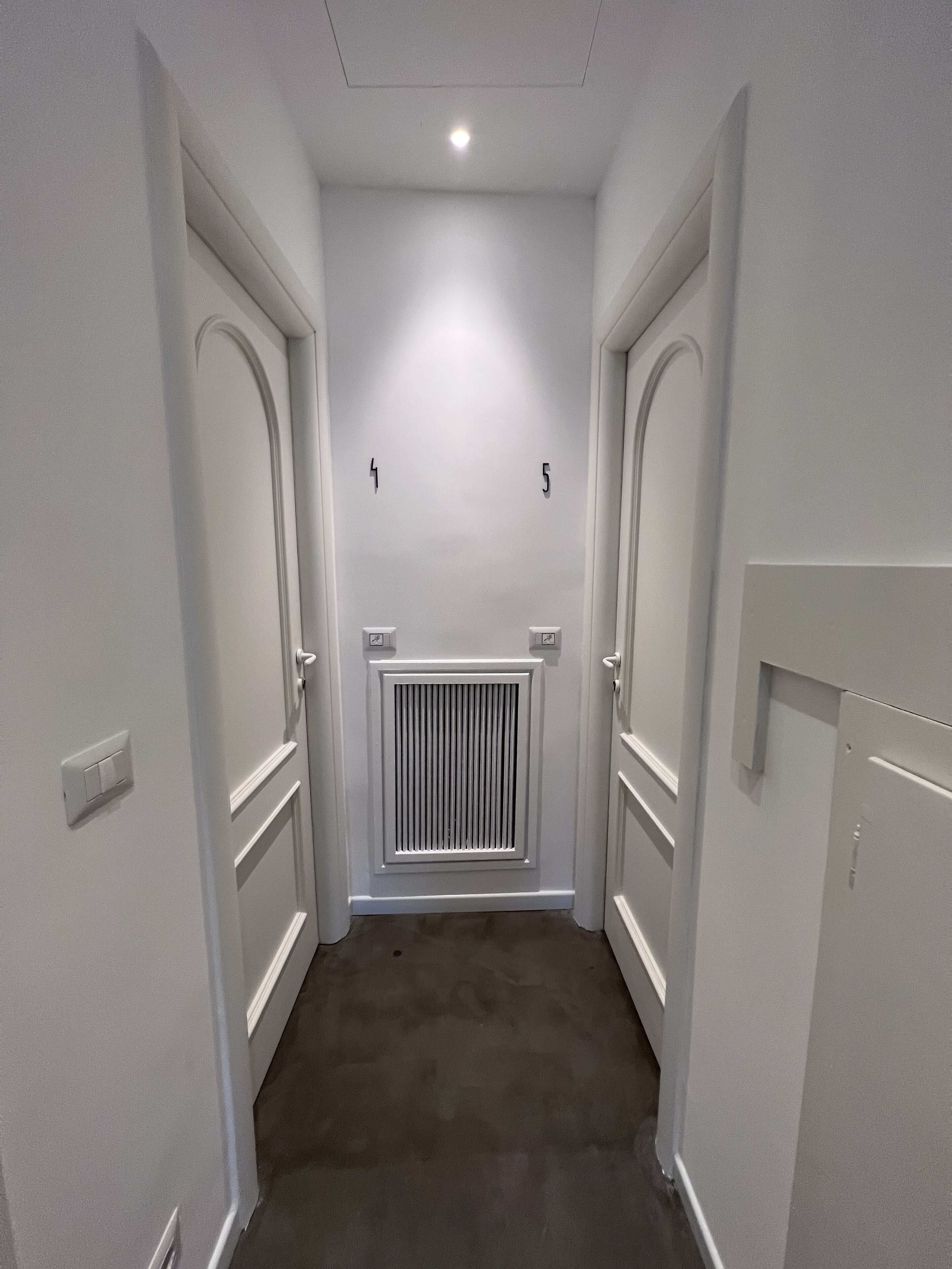 Un couloir étroit aux murs blancs et au tapis gris. De chaque côté, il y a deux portes, chacune équipée d’une sonnette et d’un interrupteur. Un radiateur est fixé au mur, entre les deux portes.