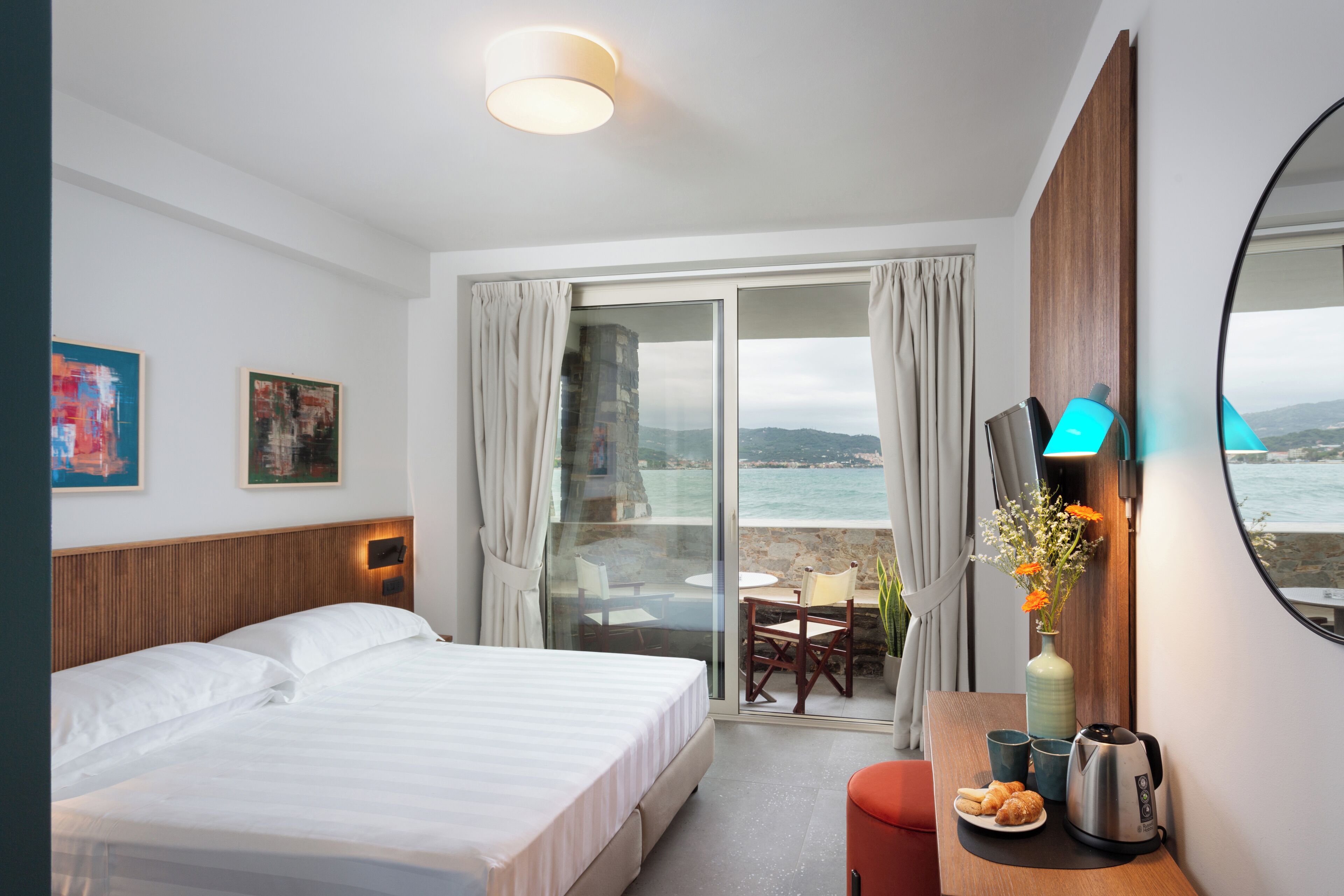 Ein Hotelzimmer mit Bett, Schreibtisch mit Kaffeemaschine, Blumen in einer Vase, Blick aufs Meer und einem Bild an der Wand.
