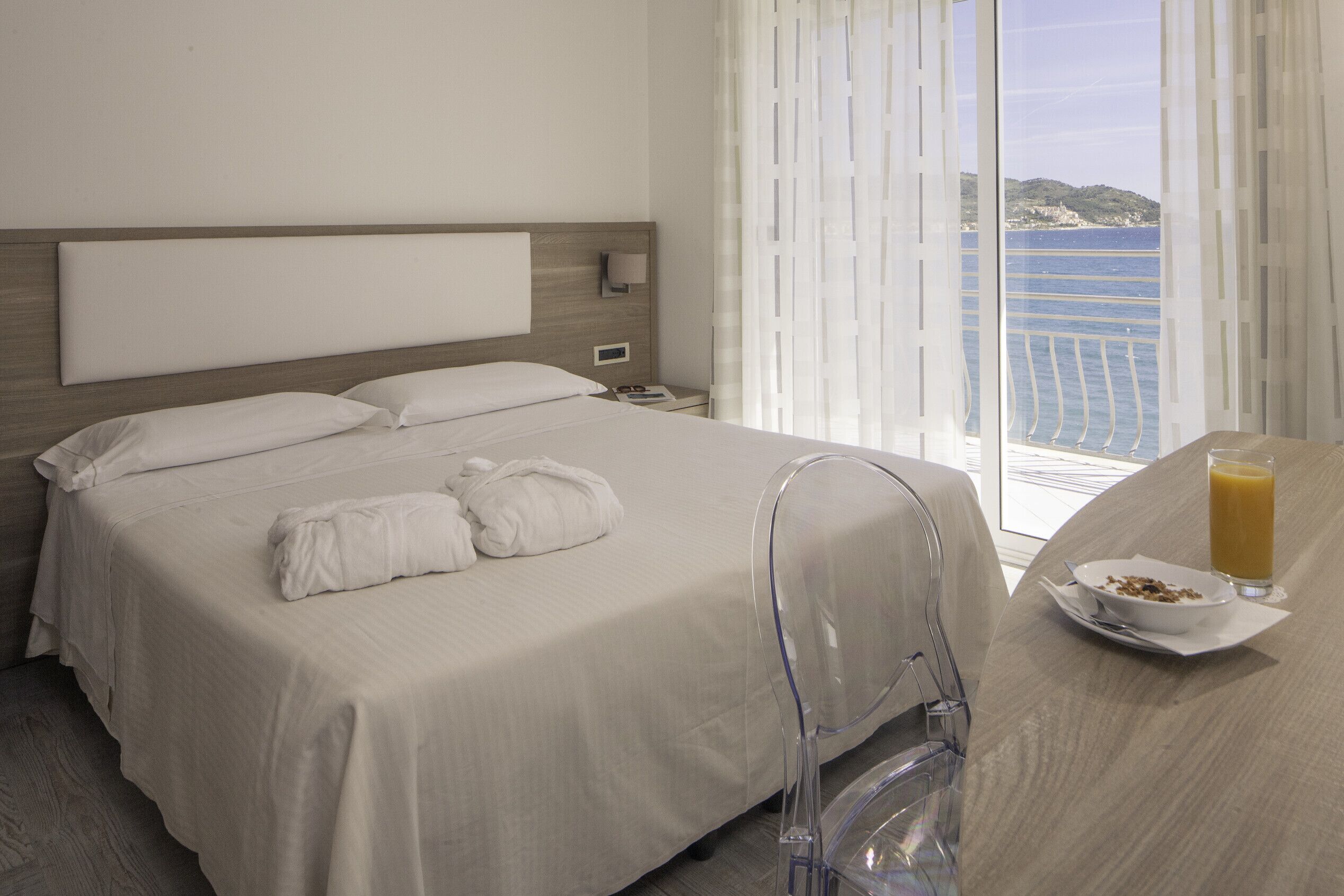 Quarto de hotel com uma cama, uma mesinha de cabeceira com um copo de suco de laranja e vista para o mar.