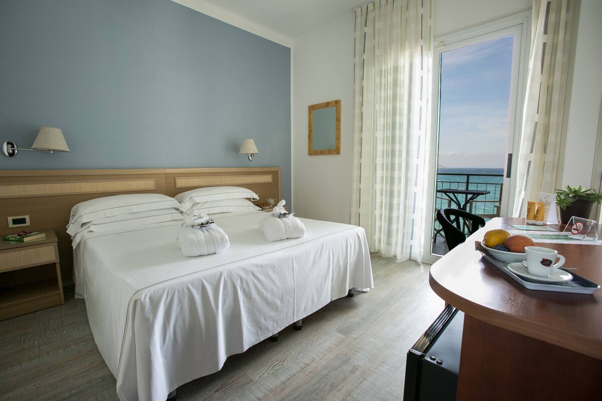 Une chambre d’hôtel avec un lit, des lampes de chevet, une petite table avec un plateau, une chaise et une vue sur la mer par la fenêtre.