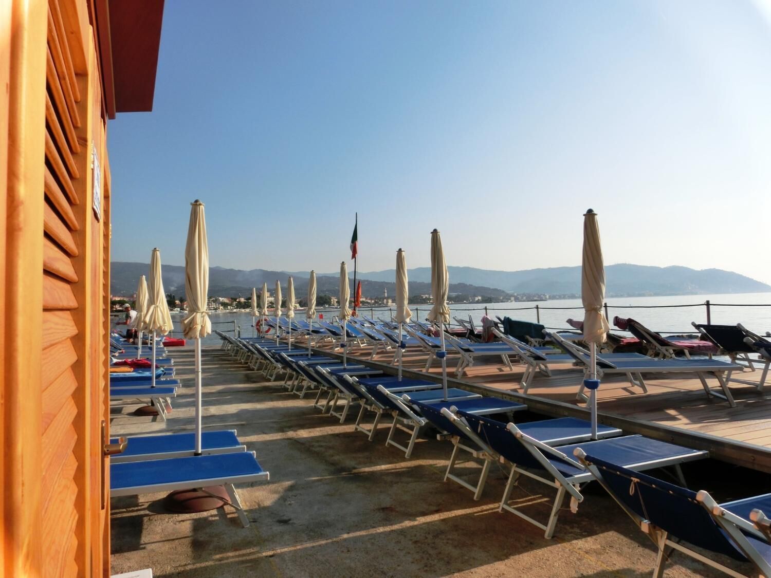 A beachside lounge area with rows of sun loungers and umbrellas, overlooking a body of water and distant mountains.