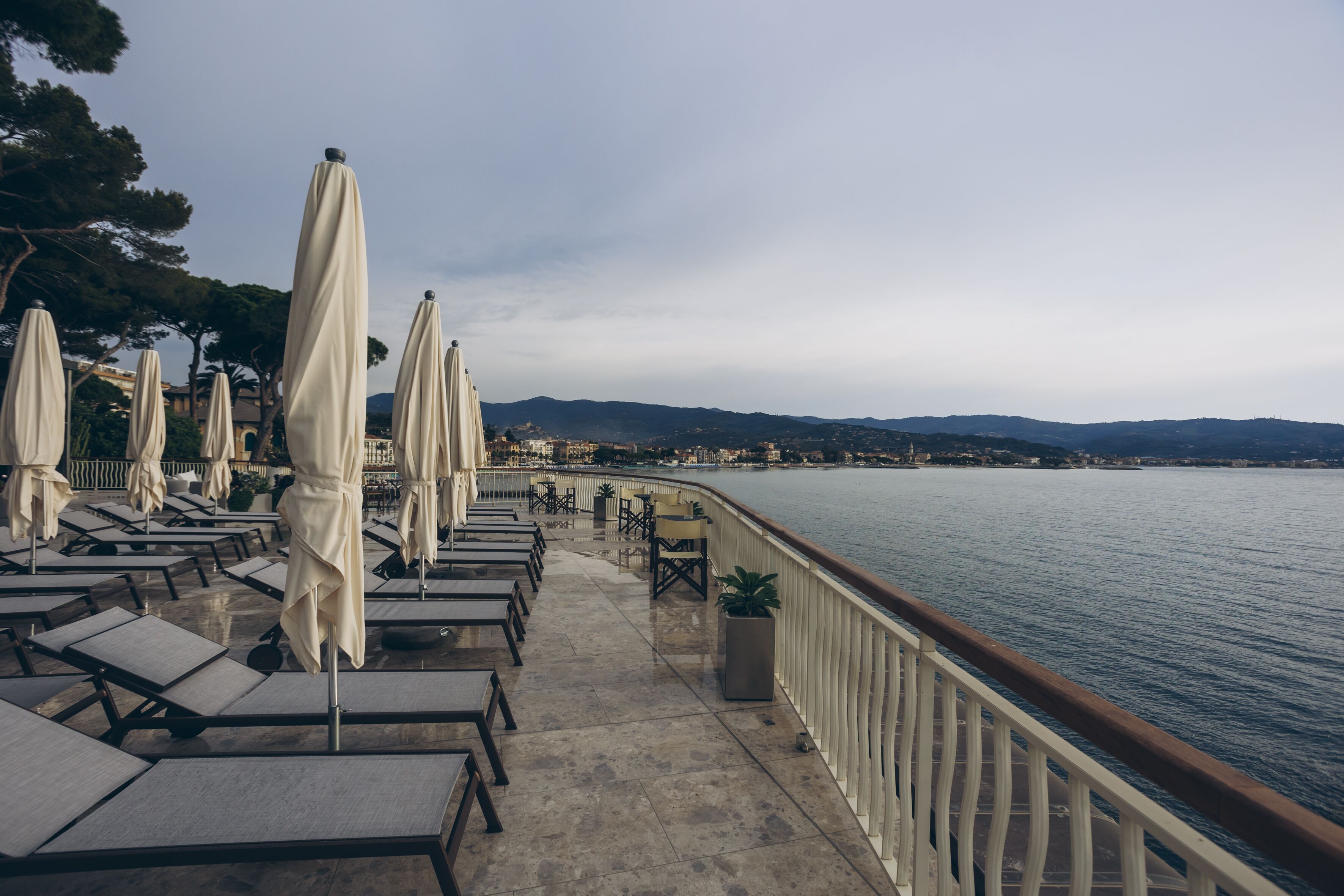 A seaside terrace with sun loungers and umbrellas, a railing, and a view of the water and distant hills.