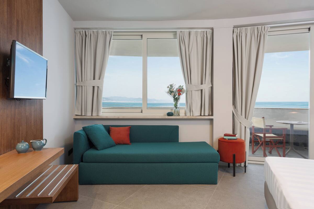 Une chambre d’hôtel moderne avec un canapé de couleur turquoise, une table d’appoint en bois, une télévision, un balcon avec vue sur la mer et une petite table avec des chaises.