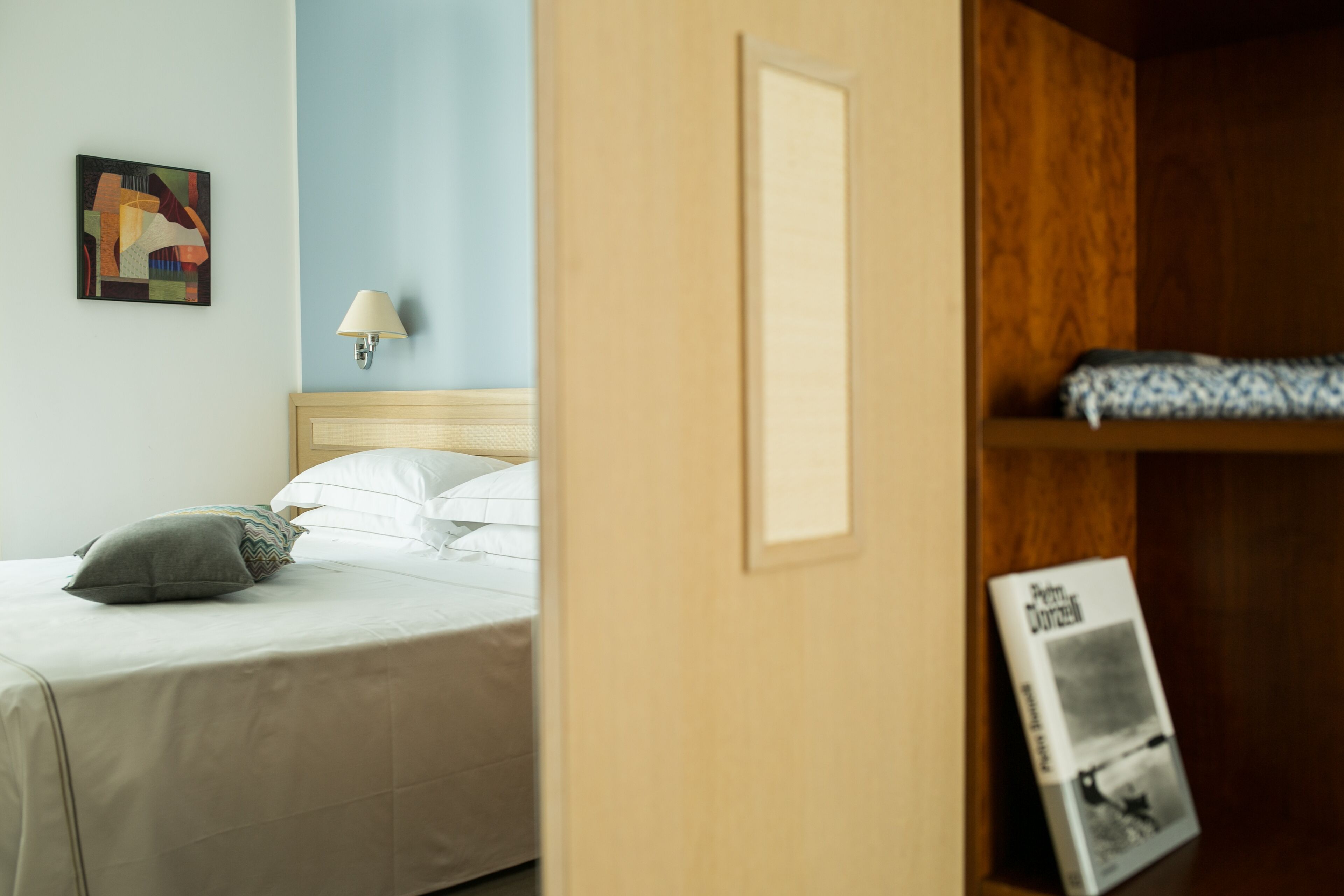 Ett hotellrum med en säng, en vägglampa, en inramad tavla och en hylla med en bok.