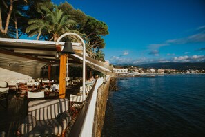 Ein Restaurant am Meer mit Außenplätzen, Blick auf das Wasser, Palmen und eine Stadt im Hintergrund.