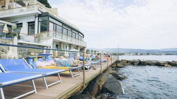Hotel frente al mar con terraza y vistas al océano, con tumbonas y fachada de piedra.