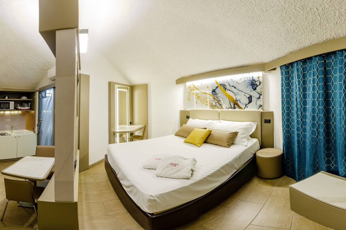 A hotel room with a bed, a desk, a chair, a small kitchenette, and a bathroom.