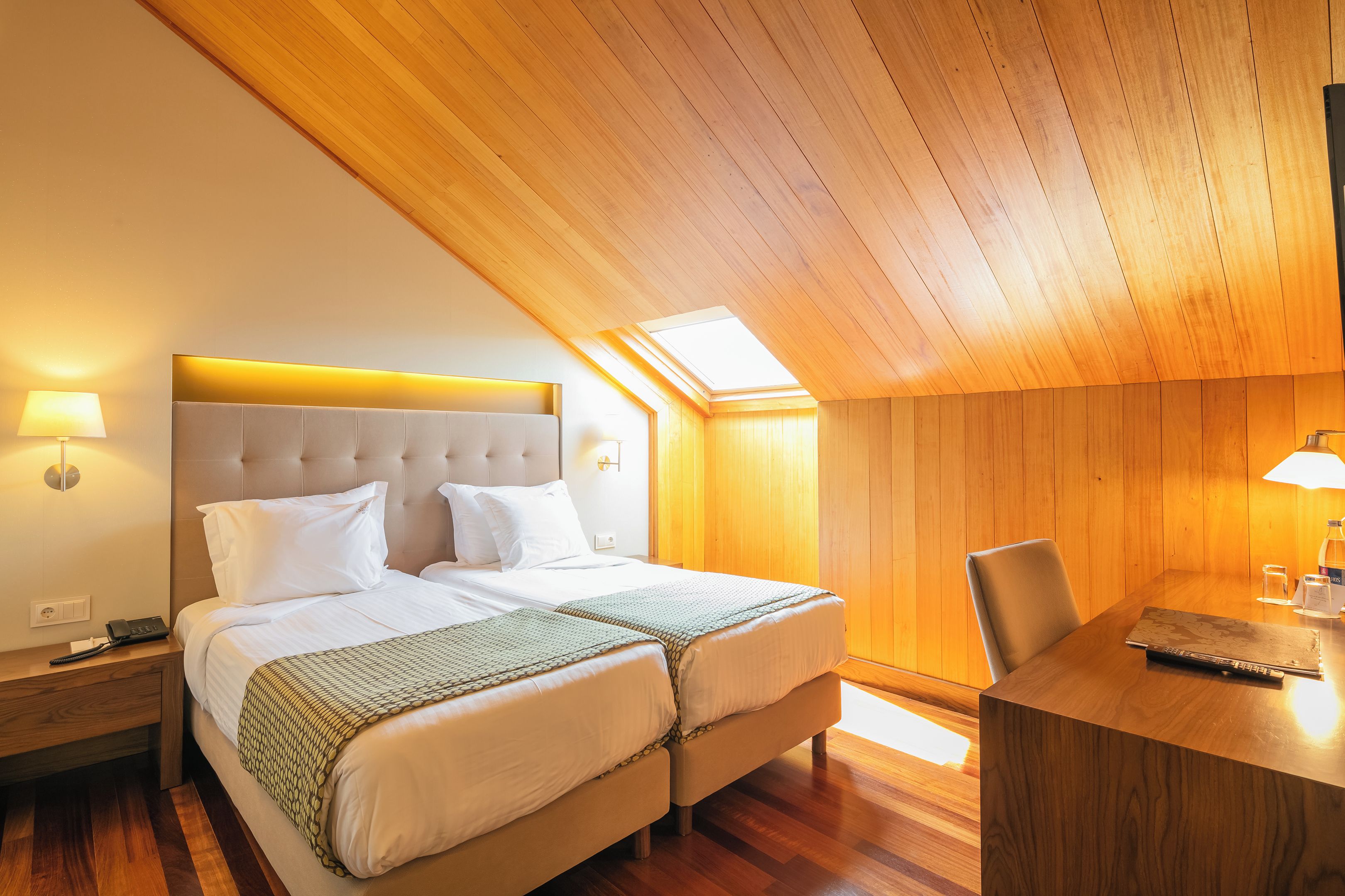 Quarto de hotel com teto de madeira, cama com lençóis brancos, escrivaninha com cadeira e uma pequena mesa com abajur.