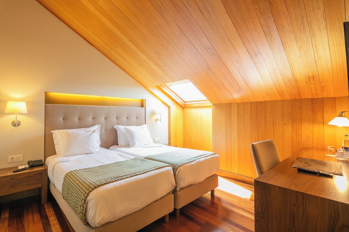 Quarto de hotel com teto de madeira, cama com lençóis brancos, escrivaninha com cadeira e uma pequena mesa com abajur.