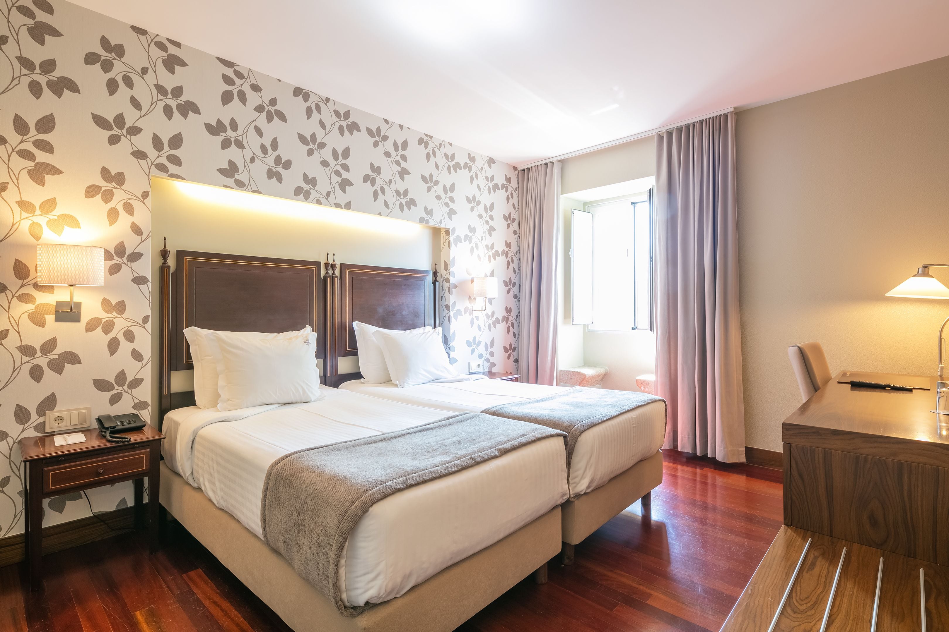 A hotel room with two beds, a desk, and a chair. There is a lamp on the desk and a lamp on the nightstand.
