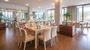A spacious dining area with multiple tables and chairs, large windows offering a view of the outdoors, and a central wooden planter with a tree.