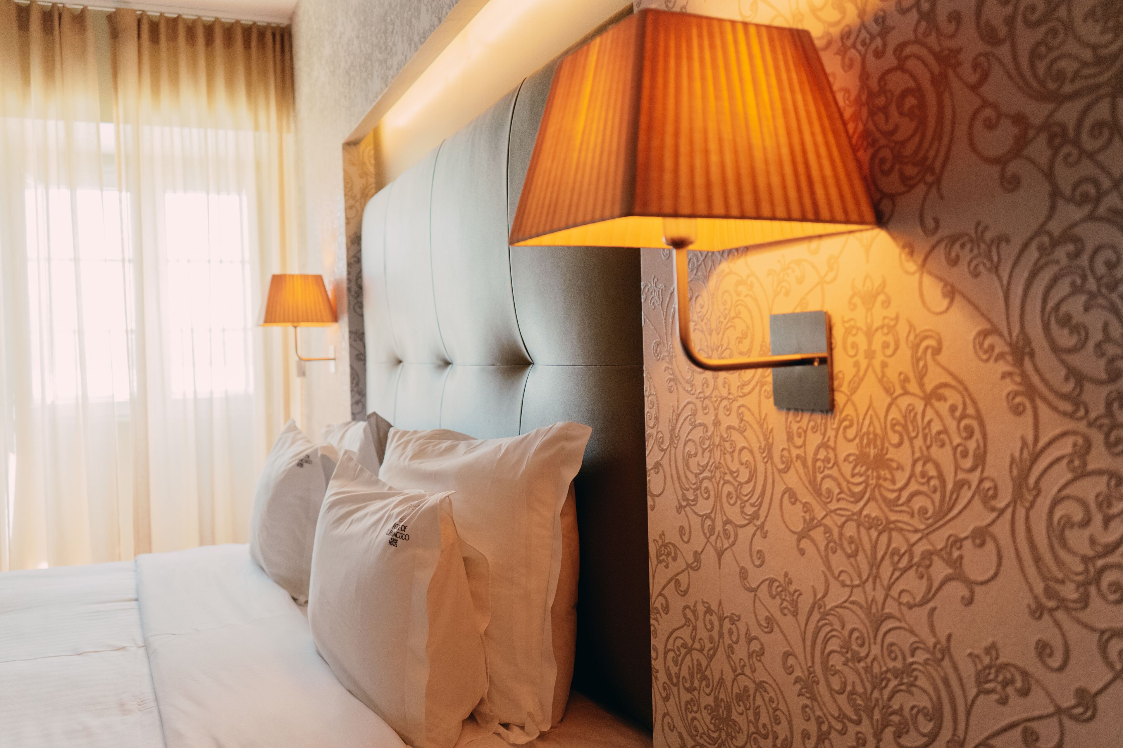 A hotel room with a tufted headboard, a bed with white linens, two bedside lamps, and a patterned wallpaper.