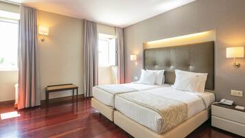 Quarto de hotel com duas camas, mesa de cabeceira e janela com cortinas.
