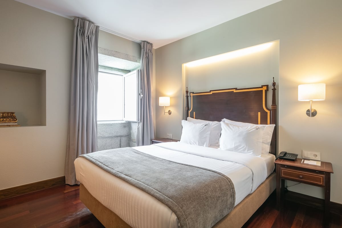 Un lit bien fait, avec une tête de lit, une table de chevet avec une lampe et une fenêtre avec des rideaux.