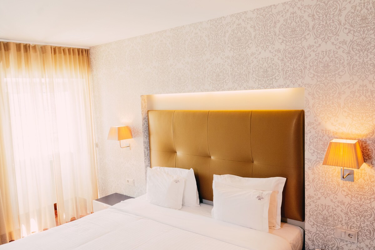 Quarto de hotel com uma cama grande, duas luminárias de cabeceira e um papel de parede estampado.