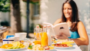 A breakfast table with various food items and drinks, a book, and a person reading.