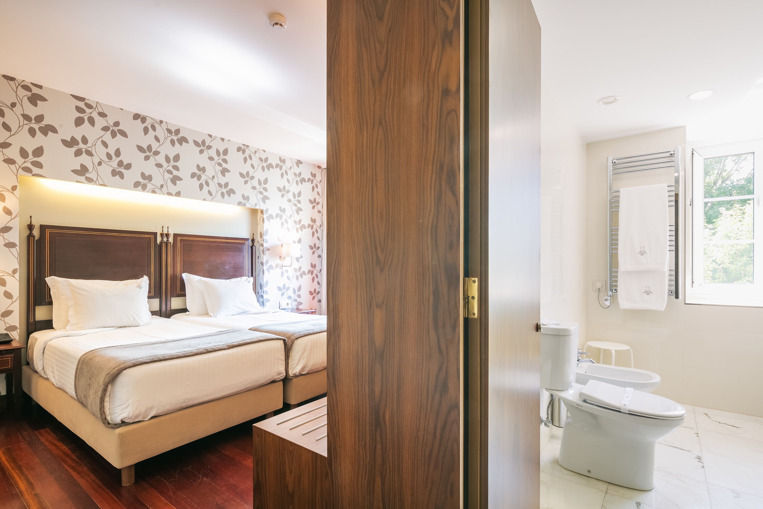 Habitación de hotel con cama, mesita de noche, puerta de madera, baño con inodoro y lavabo, y ventana.
