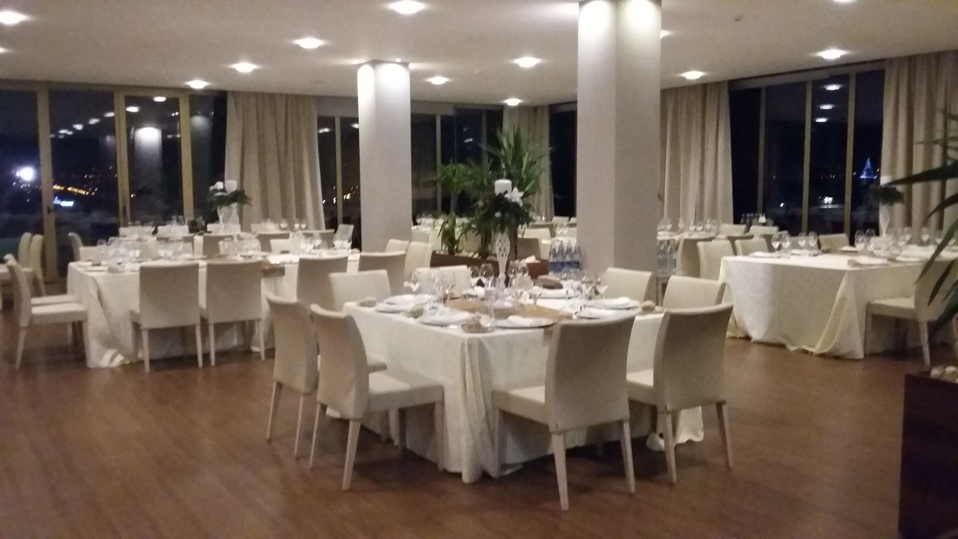 A spacious dining area with multiple round tables set for a formal event, large windows offering a view of the city at night, and a modern interior design.
