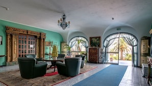 A spacious lobby with a central seating area, a chandelier, and a view of palm trees through large arched windows. - Arathena Rocks Hotel (Giardini Naxos)