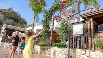 Un campo da basket all'aperto con un cartello, palme e un edificio sullo sfondo.
